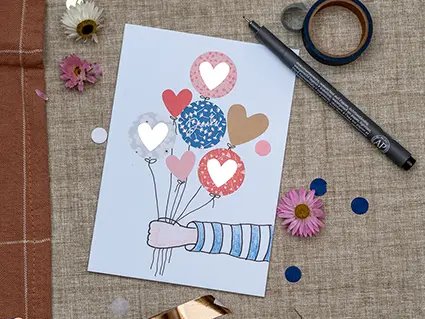 Auf einem Juteuntergrund liegt eine selbstgestaltete, weiße Klappkarte, Ein gezeichneter Arm ragt von rechts auf die Vorderseite. Der Arm hält einige Luftballons, die aus farblich aufeinander abgestimmten Aufklebern von Artoz bestehen. Um die Karte ist Konfetti und ein paar Trockenblumen verteilt. Ein schwarzer Fineliner und eine Rolle Washitape sind ebenfalls zu sehen. Foto: Johanna Rundel