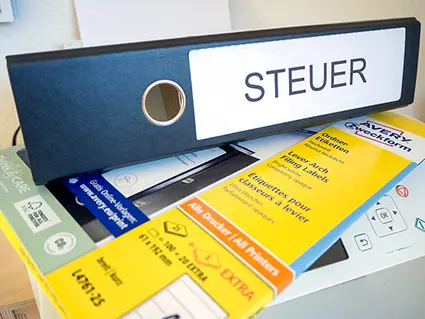 Auf einem Drucker liegt eine Packung Ordneretiketten von Avery Zweckform, darauf ein schwarzer Ordner, auf dessen Rücken „Steuer“ steht. Foto: Johanna Rundel