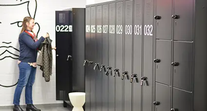 Eine Frau steht in einer Umkleide vor einer langen Schrankwand und hängt ihre Jacke in einen Spind.