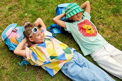 alt="Ein Mädchen und ein Junge im Grundschulalter liegen entspannt auf einer grünen Wiese. Ihre Köpfe sind auf ihren Schulranzen von ergobag gebettet. Sie haben die Hände hinter ihrem Kopf verschränkt und schauen in den Himmel. Beide tragen bunte Kleidung. Das Mädchen hat eine Sonnenbrille im Haar. Der Junge hat einen grünen Buckethat in die Stirn gezogen."