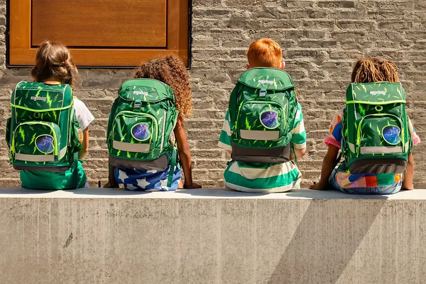 Vier Grundschüler sitzen auf einem Mäuerchen vor einem Gebäude aus grauem Klinker. Sie sind nur von hinten zu sehen und haben alle einen grünen Schulranzen von ergobag auf dem Rücken. Es sind verschiedene Modelle jedoch im gleichen Design.