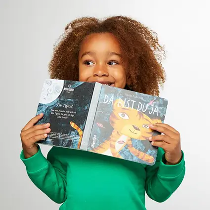 Ein Mädchen versteckt sich lachend hinter dem Papp-Bilderbuch „Da bist du ja!“ von Affenzahn.