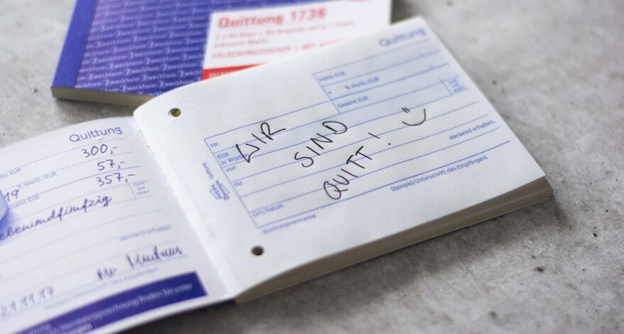 Ein Quittungsblock von Avery Zweckform liegt aufgeschlagen auf einem Tisch. Quer über die erste Seite steht der Text "Wir sind quitt" mit einem Smiley.