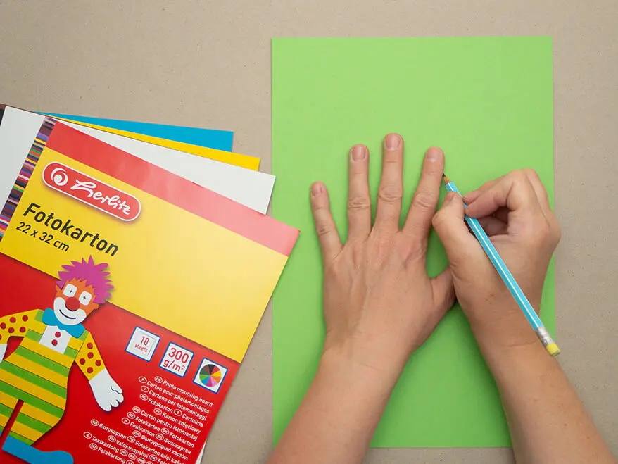 Jemand zeichnet mit Bleistift die Umrisse seiner Hand auf einen grünen Fotokarton von Herlitz ab. Foto: Johanna Rundel
