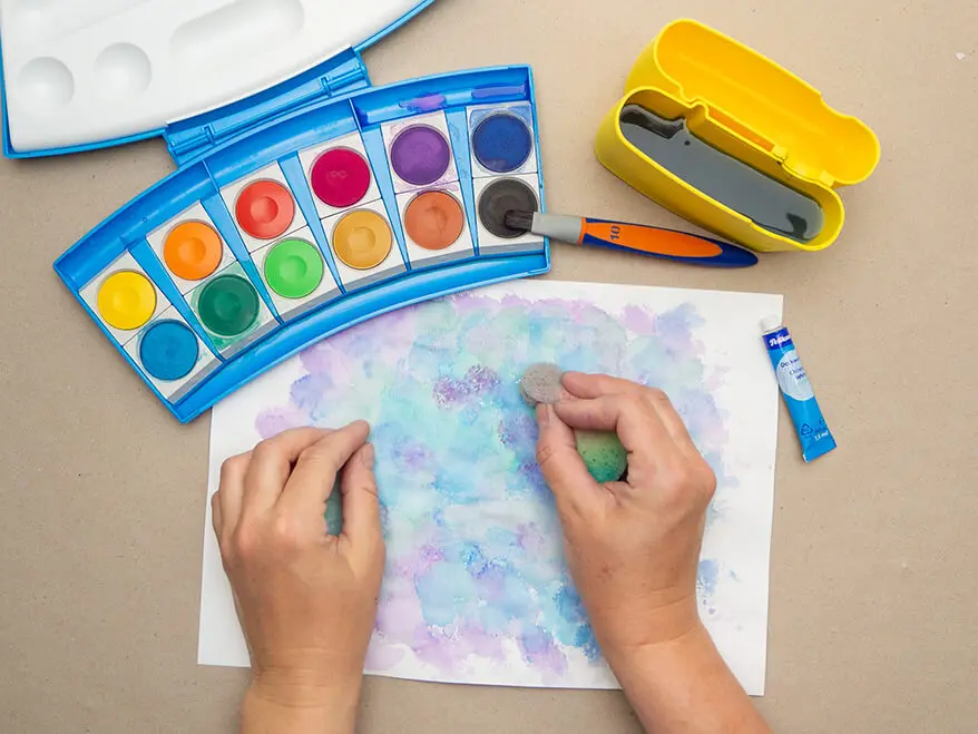 Jemand tupft mit einem Malschwämmchen Farben aus einem Wasserfarbkasten von Pelikan auf ein weißes Blatt Papier. Foto: Johanna Rundel