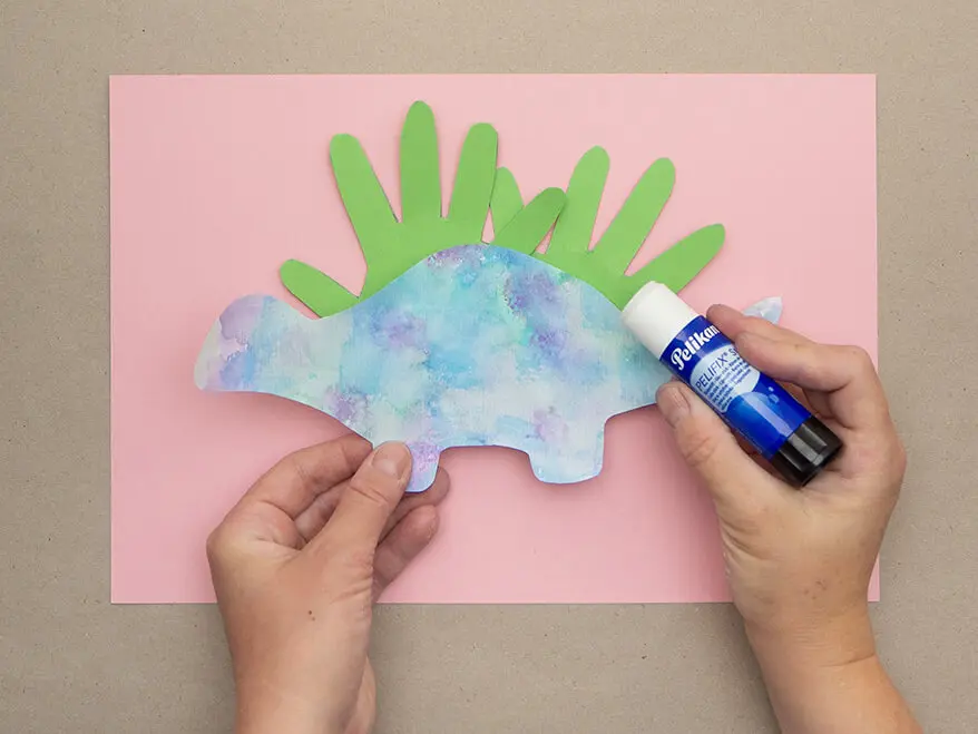 Zwei aus Fotokarton von Herlitz ausgeschnittene Handumrisse und eine Dinosaurier-Silhouette aus Papier werden mit einem Klebestift von Pelikan auf ein rosa Papier geklebt. Foto: Johanna Rundel