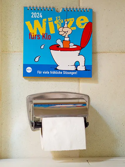 Über einem silbernen Klopapier-Halter an einer hellgrün gekachelten Wand hängt ein "Witze fürs Klo"-Kalender des Athesia Kalenderverlags. Foto: Johanna Rundel