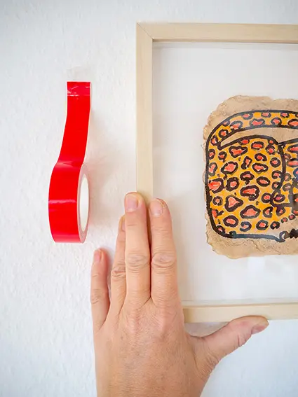 Jemand hängt an einer weißen Wand ein Bilderrahmen auf. Daneben klebt eine rote Rolle Spezialklebeband „Kleben statt bohren“. Foto: Johanna Rundel
