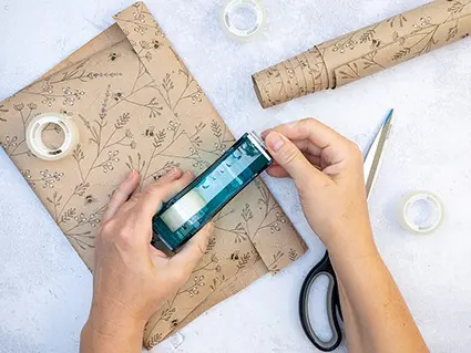 Jemand packt mit transparentem Klebefilm ein Geschenk in Packpapier ein. Dafür nutzt er einen Klebebandabroller. Foto: Johanna Rundel