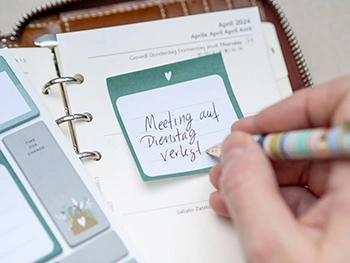 Jemand schreibt mit einem pastellfarbenen Kugelschreiber „Meeting auf Dienstag verlegt“ auf einen Klebezettel in einem Filofax-Planer. Foto: Johanna Rundel 