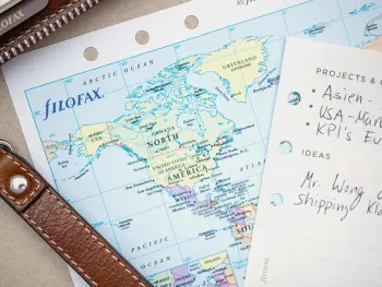 Eine aufgeheftete Weltkarte liegt neben einem braunen Filofax-Planer aus Leder. Darauf ein Zettel mit Tod-Dos. Foto: Johanna Rundel 