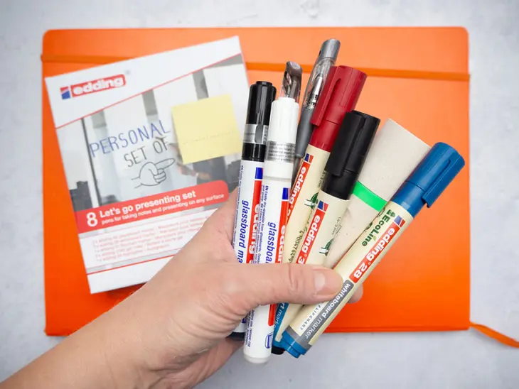 Jemand hält verschiedene Stifte von edding vor eine Schachtel, auf der „Personal Set of…“ steht. Darunter liegt ein orangenes Notizbuch. Foto: Johanna Rundel