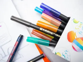 Viele bunten Brushpens von edding ergießen sich aus einer Pappschachtel auf eine Skizze. Foto: Johanna Rundel