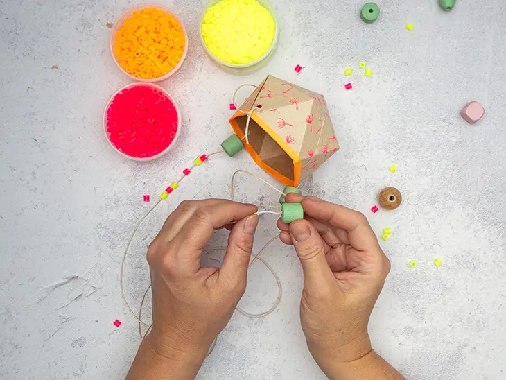 Jemand fädelt bunte Perlen von KNORR prandell auf die Schnur einer selbstgebastelten Blumenampel aus einer upcycelten Milchtüte.