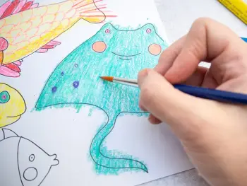 Jemand aquarelliert mit einem Pinsel einen mit Stiften von STAEDTLER ausgemalten Fisch für ein DIY-Aquarium aus Karton. Foto: Johanna Rundel