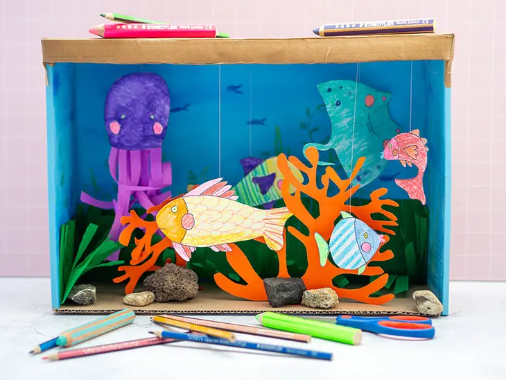 Ein selbstgebasteltes Aquarium aus einem Pappkarton steht vor einer rosa Wand. Davor und darauf liegen Stifte von STAEDTLER. Foto: Johanna Rundel