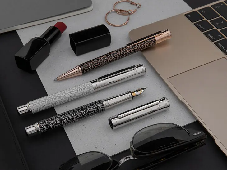 Schreibgeräte in verschiedenen Metallic-Designs liegen auf einem femininen Arbeitsplatz zwischen Laptop, Lippenstift und Sonnenbrille.