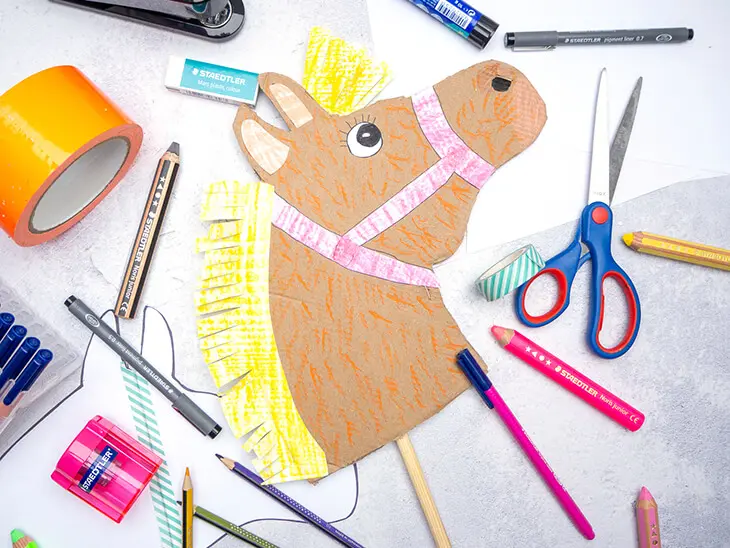 Ein selbstgebasteltes Steckenpferd aus Pappe liegt zwischen vielen verschiedenen Bastelmaterialien von STAEDTLER auf einem hellgrauen Untergrund. Foto: Johanna Rundel