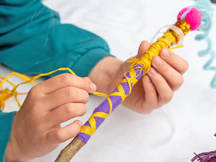 Ein Grundschulkind wickelt eine gelbe Bastschnur von HEYDA um einen selbstgebastelten Zauberstab aus einem Stock. Foto: Johanna Rundel