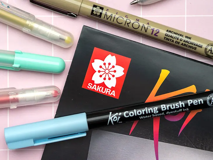 Verschiedene Stifte und Papier von Sakura liegen verstreut auf einem rosa Untergrund. Foto: Johanna Rundel