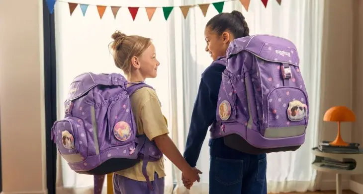 Zwei Mädchen mit lila Schulranzen von ergobag halten sich an der Hand und lächeln sich an