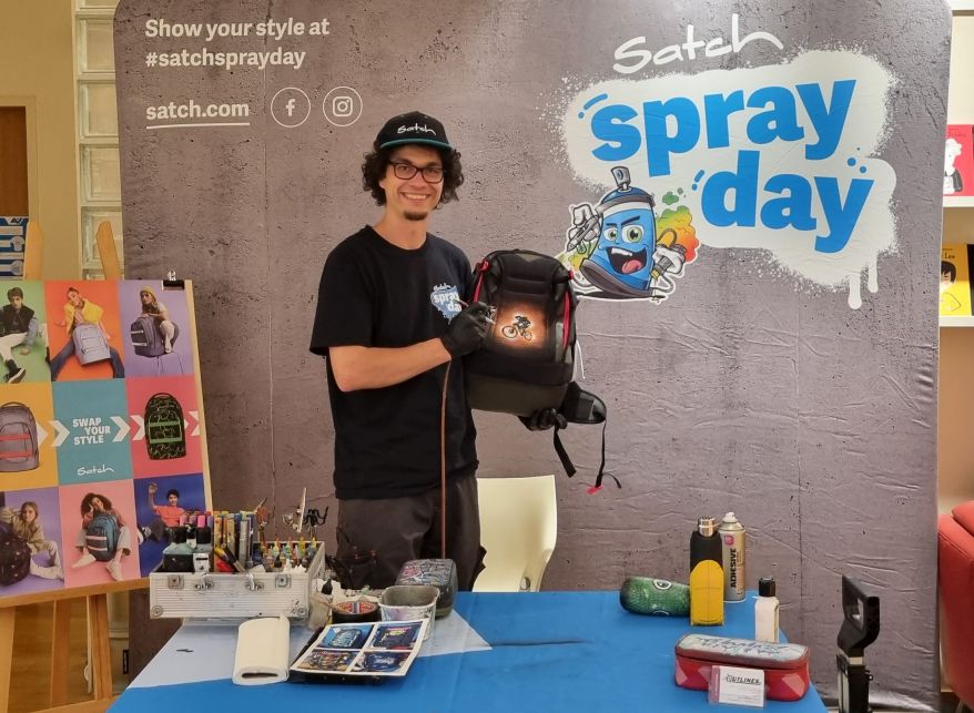Ein Grafittikünstler steht vor einem Plakat für den Satch Spray Day. Er hält lächelnd einen Schulrucksack der Marke satch in die Luft, den er mit einem Grafitti verziert hat. Vor ihm stehen auf einem Tisch diverse Farbdosen und Kreativmaterial.
