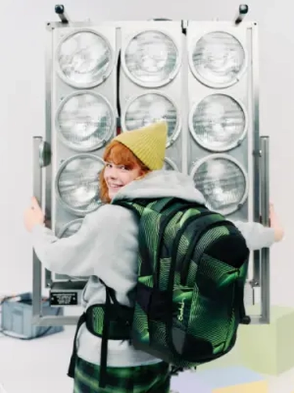 Ein Mädchen mit gelber Mütze und grün-schwarzem satch pack Rucksack steht vor einem Scheinwerfer und dreht sich um zur Kamera.