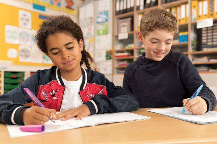 2 Kinder sitzen an einem Tisch und schreiben etwas in ihre Schulhefte. 