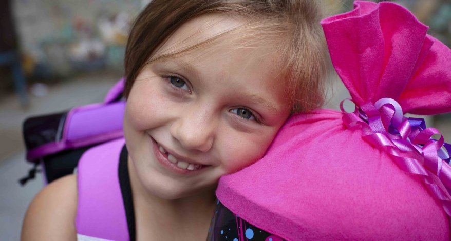 Ein Mädchen mit einem lila Schulranzen auf dem Rücken hält eine Schultüte mit pinkem Verschluss in der Hand. Sie lehnt den Kopf an die Schultüte und lächelt in die Kamera.
