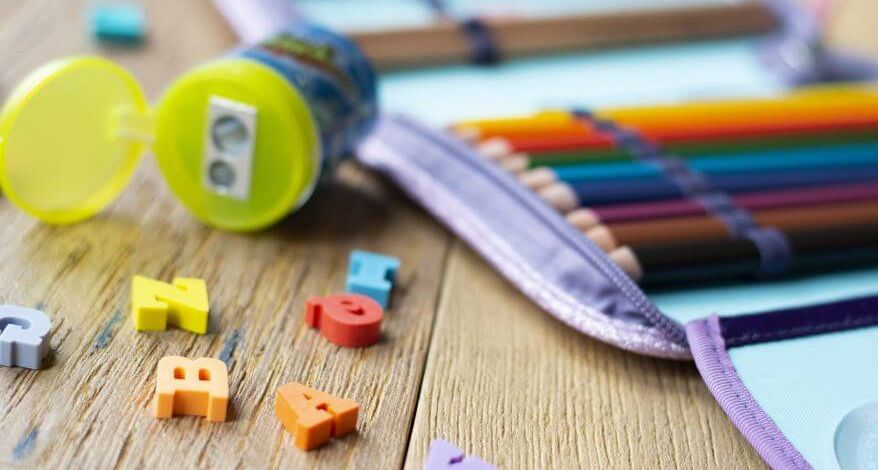 Auf einem Holztisch liegt ein aufgeschlagenes, mit Buntstiften befülltes Federmäppchen. Daneben liegt eine geöffnete Spitzerdose und kleine bunte Radiergummi-Buchstaben.