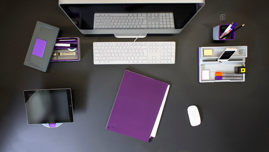 Bild aus der Vogelperspektive auf einen schwarzen Tisch. Auf diesem steht ein Monitor mit einer weißen Tastatur, rechts daneben steht ein Desk Organizer mit Stiften, Spitzer und einem Smartphone, dahinter ein Stifteköcher mit Stiften. Links neben dem Monitor liegt eine geöffnete Stiftschale mit Stiften, davor steht ein Tablethalter mit einem Tablet. Vor der Tastatur liegen eine lila Kladde und eine weiße PC-Maus.