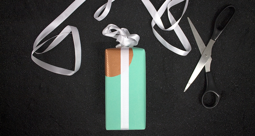 Auf einem schwarzen Untergrund liegt ein Geschenk, welches in mintgrünes Papier eingepackt ist und einen goldenen Kreis an der oberen Ecke hat. Es ist mit weißem Schleifenband verpackt. Ein Rest des Schleifenbands und eine Schere liegen neben dem Geschenk.