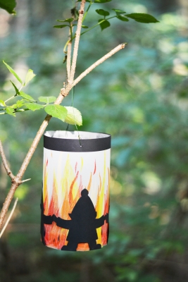 An einem Ast hängt eine selbstgebastelte Sankt Martins Laterne mit kleinen Wichteln, die ums Feuer tanzen.