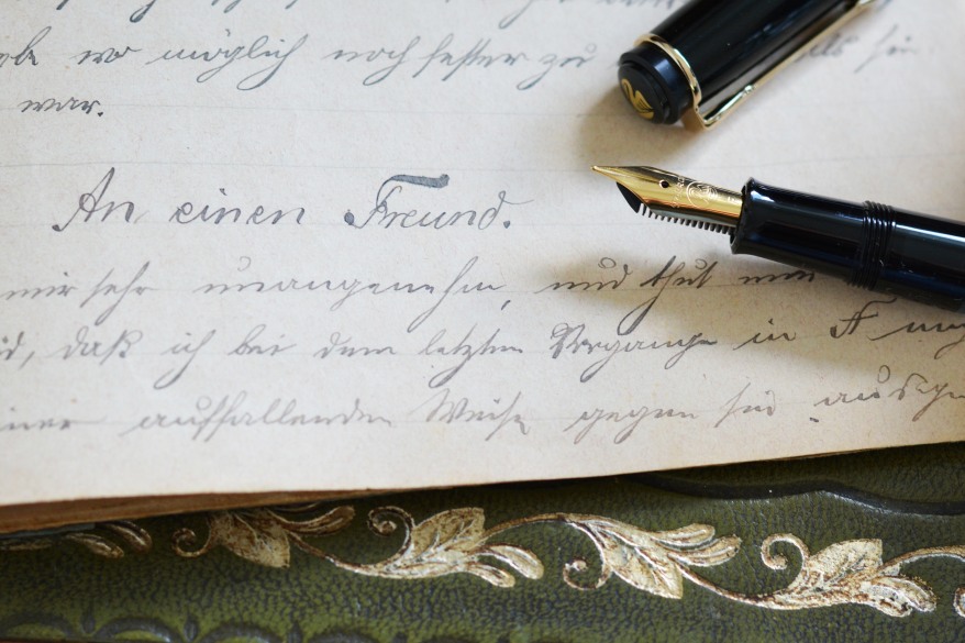 Auf einem grünen Untergrund mit hellem Blumenmuster liegt ein altes Schriftstück. Auf diesem ist in Schreibschrift der Satz "An einen Freund" zu lesen. Im rechten Bildrand liegt ein geöffneter Füller in schwarz der Marke Pelikan. Die Feder und der Clip an der Füllerkappe sind gold.  