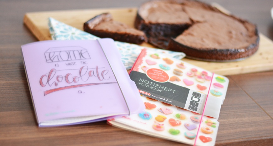 Auf einem Holztisch liegen zwei Notizhefte der my.book flex Reihe von Herlitz. Das linke Buch wurde mit den Worten "Home is where the chocolate is" belettert. Im Hintergrund ist ein angeschnittener Schokolagenkuchen auf einem Holzbrett zu sehen.