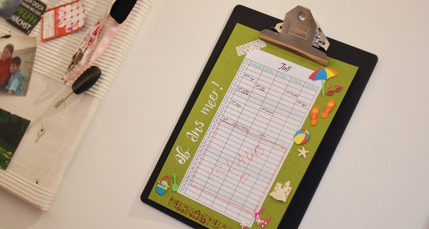 Bunter Urlaubsplan für Juli auf grünem Papier mit Clip-Halter an der Wand.