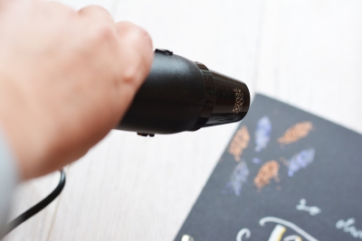 Hand nutzt Heißluftföhn, um schimmernde Federmotive auf Papier zu fixieren