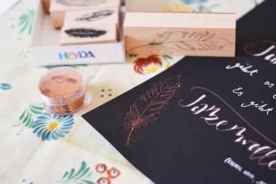 Kupferfarbene Feder auf schwarzem Papier, daneben Stempelset und Glitzerpulver von Heyda.