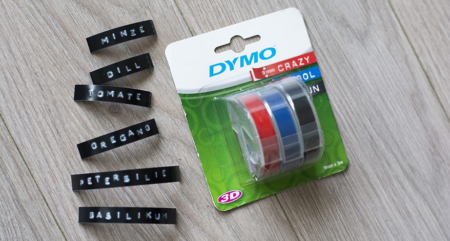 Auf einem weißen Parkettboden liegt eine Packung mit roten, blauen und schwarzen Prägeband von Dymo. Daneben liegen einige beschriftete Prägebänder mit verschiedenen Namen von Kräutern.