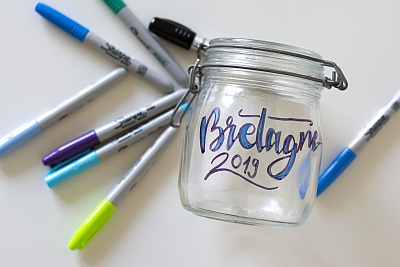 Ein Weckglas mit dem in blau geletterten Schriftzug "Bretagne 2019" liegt auf einem Tisch. Daneben liegen Permanentmarker der Marke Sharpie in unterschiedlichen Blautönen, Grün und Lila.