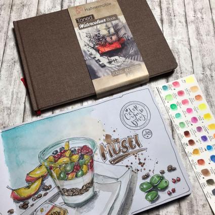 Auf einem Holzboden liegen ein geschlossenes Toned Watercolour Book von Hahnemühle in schwarz. Darunter liegt ein geöffnetes Buch mit weißes Papier, in das mit Aquarellfarben ein Glas mit Müsli und verschiedene Früchte gemalt wurden. Rechts davon liegt ein Blatt mit verschiedenen Farbproben von Aquarell-Farben. 