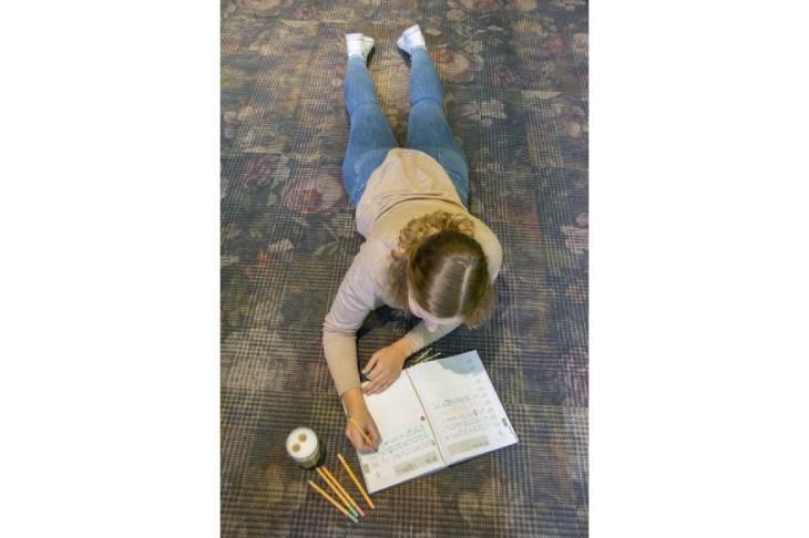 Eine junge Frau liegt mit dem 10-Jahreskalender auf dem Boden und schreibt etwas hinein. Neben dem Kalender liegen 4 bunte Fineliner und es steht ein Cappuccino daneben.