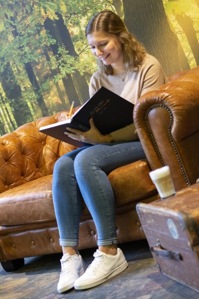 Eine junge FRau sitzt in einem Zimmer mit einer Wald-Tapete auf einer Leder-Couch. In der Hand hält sie einen 10 Jahreskalender, in den sie etwas reinschreibt.