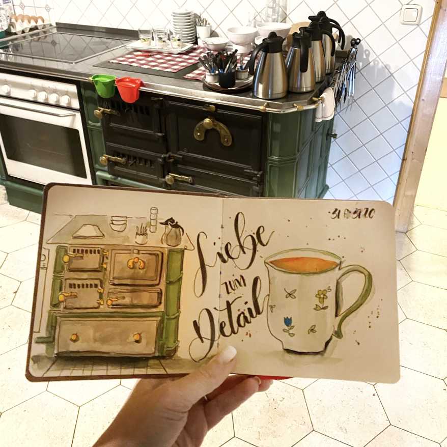 In einer Küche mit einem alten Herd hält eine Hand ein Skizzenbuch mit dem Motiv des Herds in der Hand.