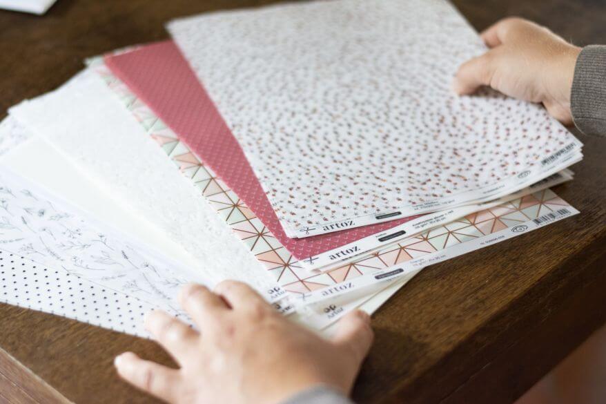 Person hält verschiedene gemusterte Bastelpapiere auf einem Holztisch.