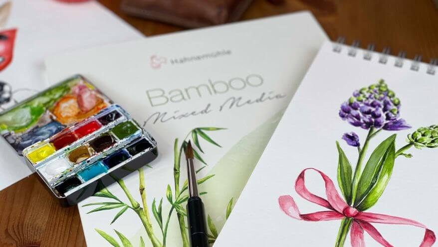 Ein Malblock Bamboo-Mix-Media-Papier aus der Serie Natural Line von Hahnemühle liegt auf einem Tisch, daneben ein aufgeklappter Farbkasten mit Aquarellfarben und ein Skizzenblock mit einer mit Aquarellfarben gemalten Blume. Foto und Illustration von Verena Knabe.