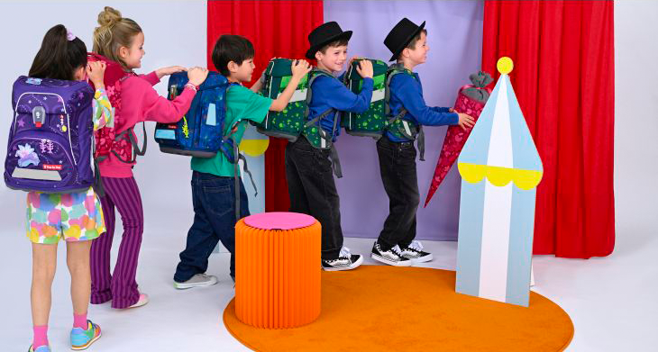 5 Kinder mit Schulranzen von Step by Step auf dem Rücken machen eine Polonaise vor einer Zirkuskulisse