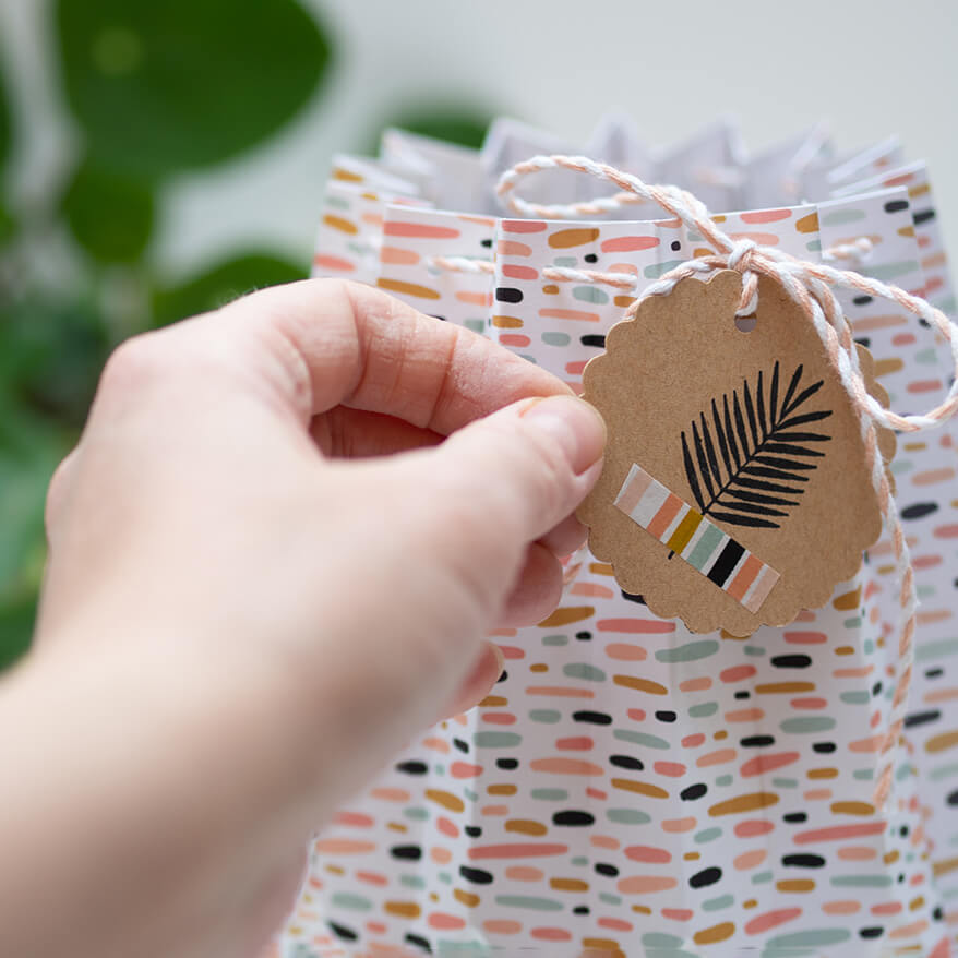 Eine Hand hält ein rundes Etikett aus Kraftpapier mit den Fingern fest. Es ist mit Bäckergarn an einer Pappvase aus Designpapier von Artoz befestigt.
