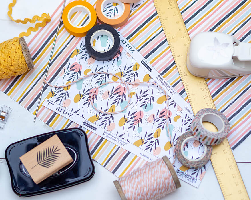 Auf einem hellen Untergrund liegen Papiere, Washitapes, Garne, Stanzer und Stempel der Marke Artoz verteilt. Daraus soll eine Pappvase gebastelt werden.