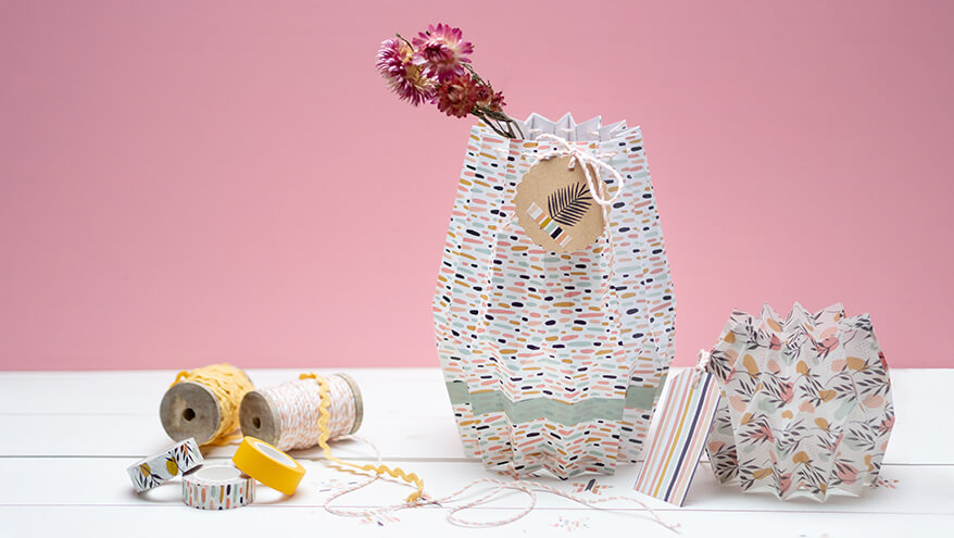 Eine gefaltete Vase und ein Teelichthalter aus Pappe mit Papieren von Artoz. In der Vase befindet sich eine rote Blume. Am linken Bildrand erkennt man bunte Washitapes und Garne der Marke Artoz. 